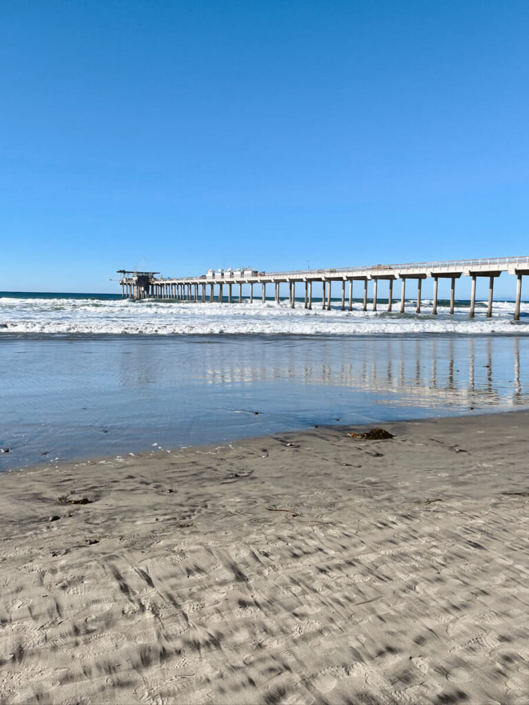 Looking out at the pier at La Jolla Shores Beach from the sand on a sunny day. There are calm waves in the ocean. 