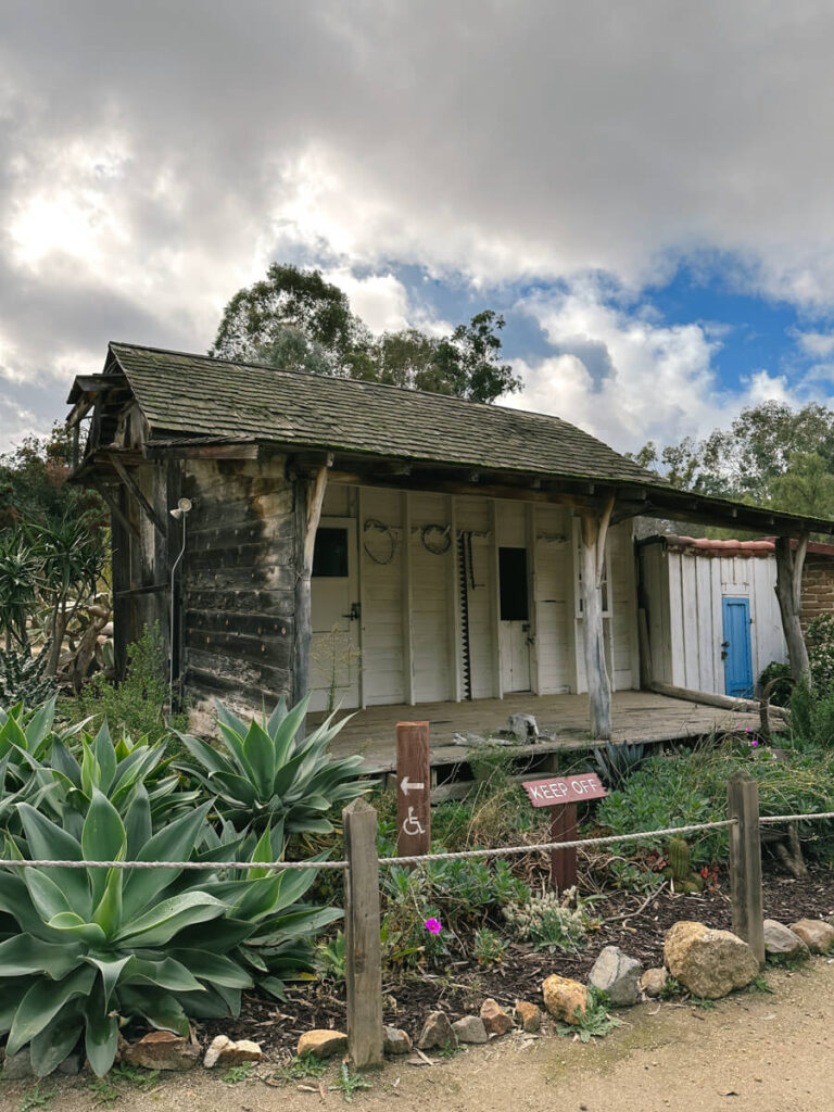 An old building with a wooden covered patio in front. There are a variety of plants in front of the building, as well as a small sign that reads "Keep Off"