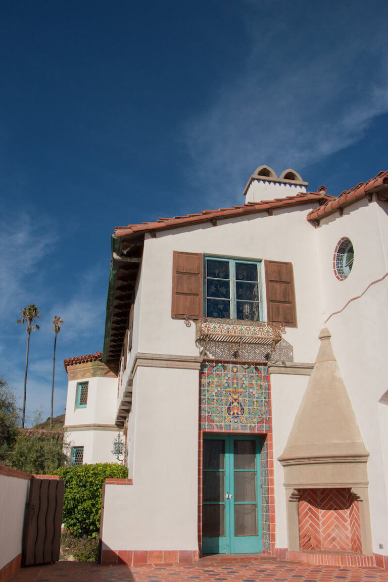 The view of a courtyard on the exterior of the Adamson House, which features red tile on the floor and teal and red decorative tile on the house.