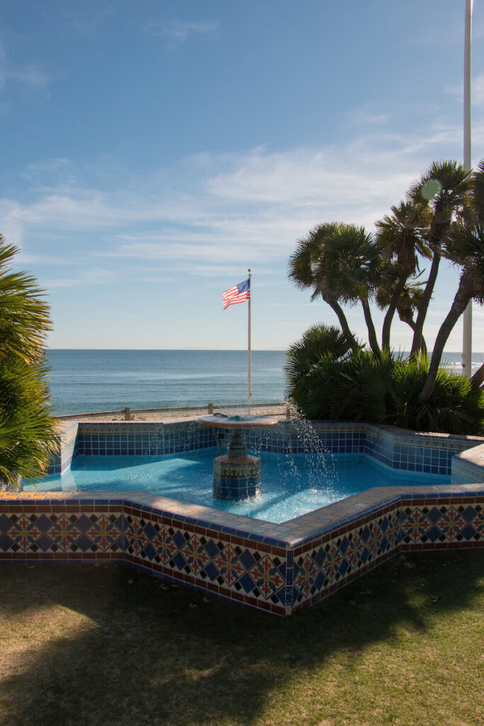 The Star Fountain at the Adamson House, with the ocean and American flag behind it. There are palm trees surrounding the fountain.