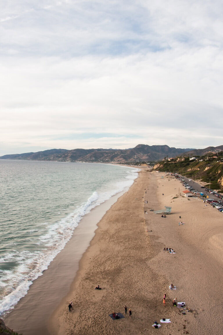 View of the Malibu Coastline during sunset, with a large open sandy space. There is the ocean to the left, and the Santa Monica Mountains to the right.