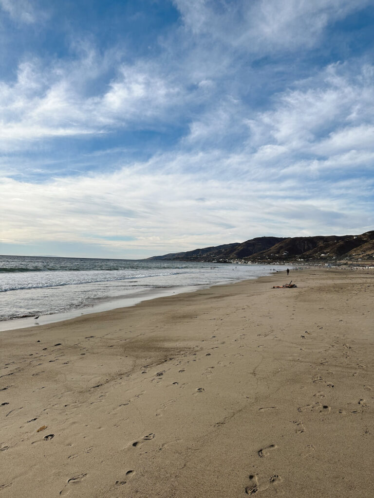 View of Zuma Beach, with the mountains in the background. The sand is to the right, and the ocean is to the left.