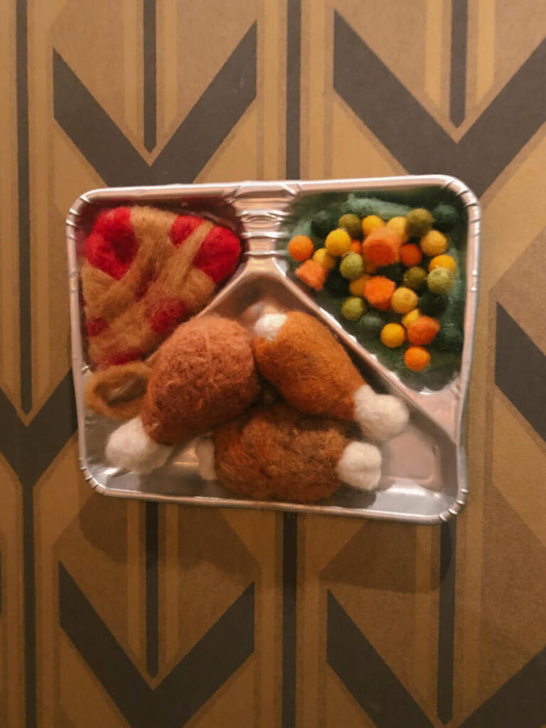 A quirky TV dinner tray made of felt, featuring chicken and a variety of chopped up vegetables. It hangs on the wall at Modern Times.