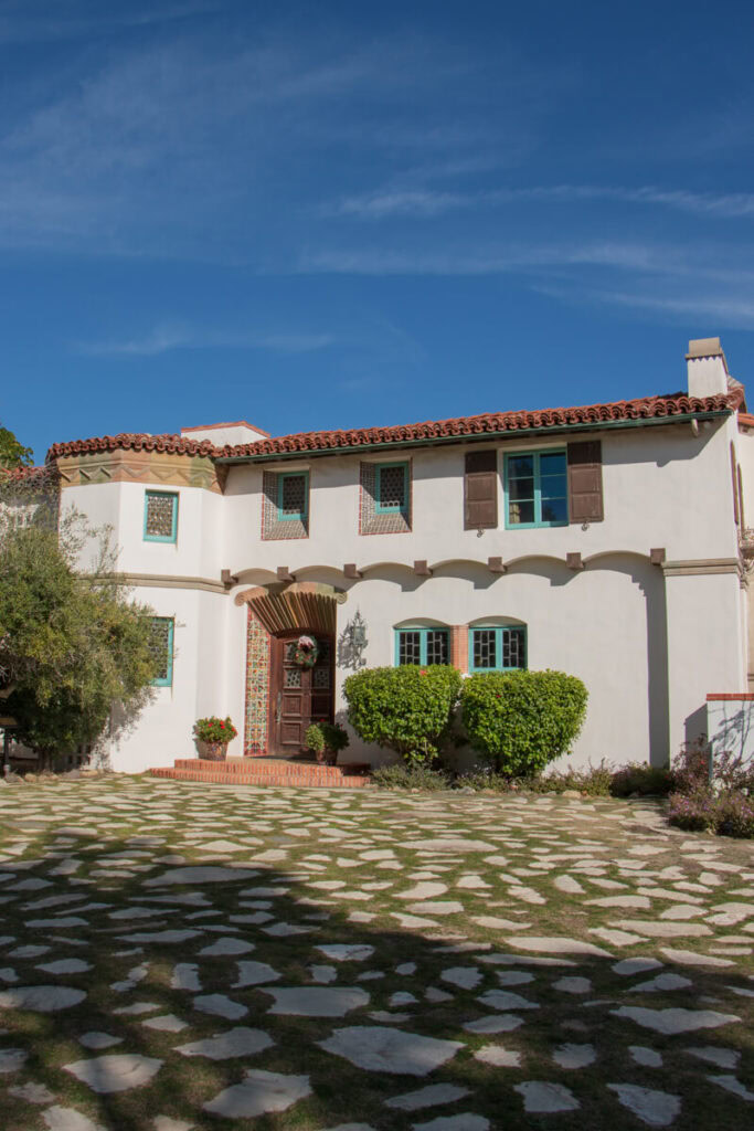 The front courtyard of the Adamson House. There are sporadic stones layed in grass on the driveway, and you can see the intricately detailed wooden front door, as well as the teal colored window sills.