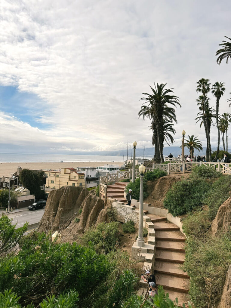 Steps from Palisades Park leading down to a pedestrian bridge. There is a portion of the road on the right, with beach houses lining the road. The ocean is in the background.