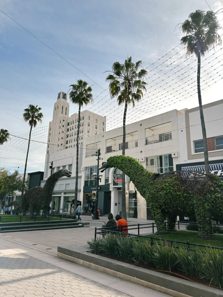 Two dinosaur shaped bushes facing each other at Third Street Promenade. There is a row of palm trees lining the walkway in the background