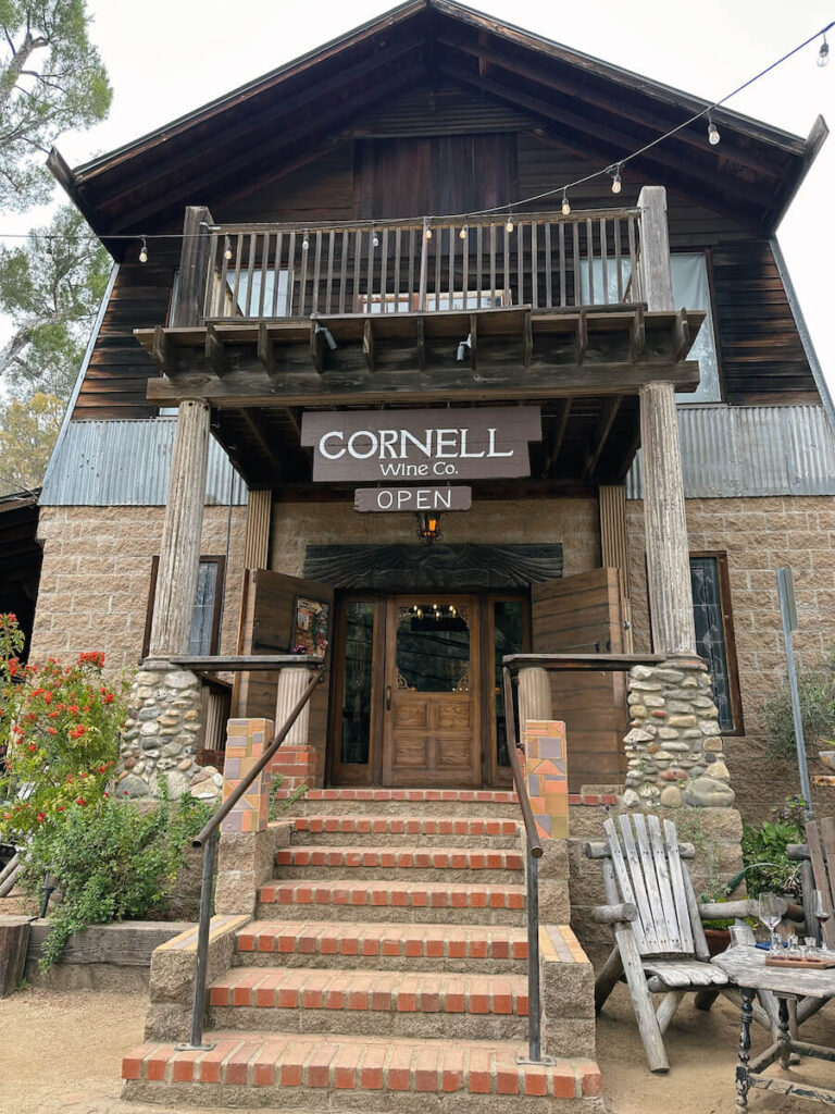 The exterior of Cornell Wine Co. There are steps leading up to a wooden door. Above the entrance, there is a hanging sign that says "Cornell Wine Co", and below that another sign says "open"