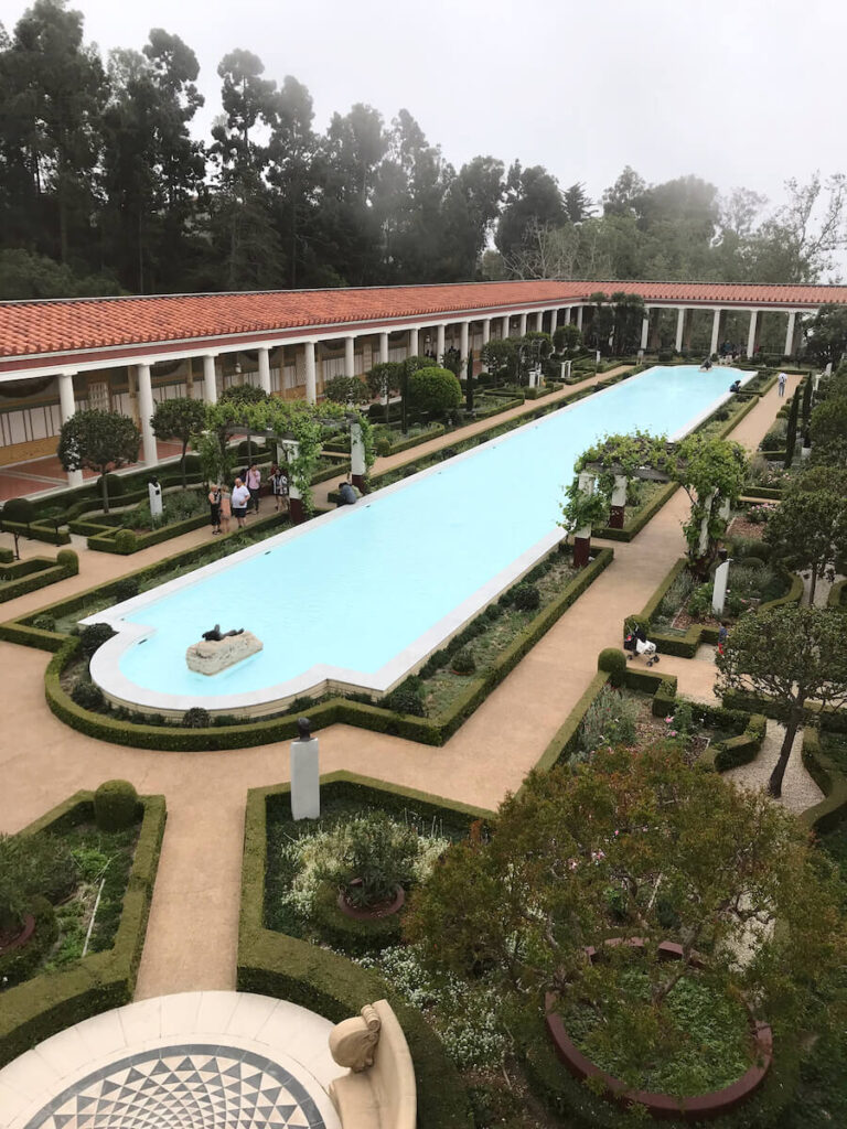 View of a outdoor water feature at The Getty Villa. The water feature is surrounded by plants an outdoor pathway that is lined with columns