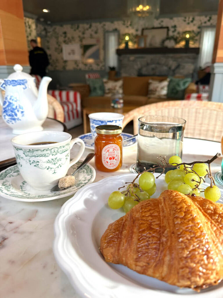 A croissant and a bunch of green grapes on a plate, with a tea kettle and two tea cups behind it on a table