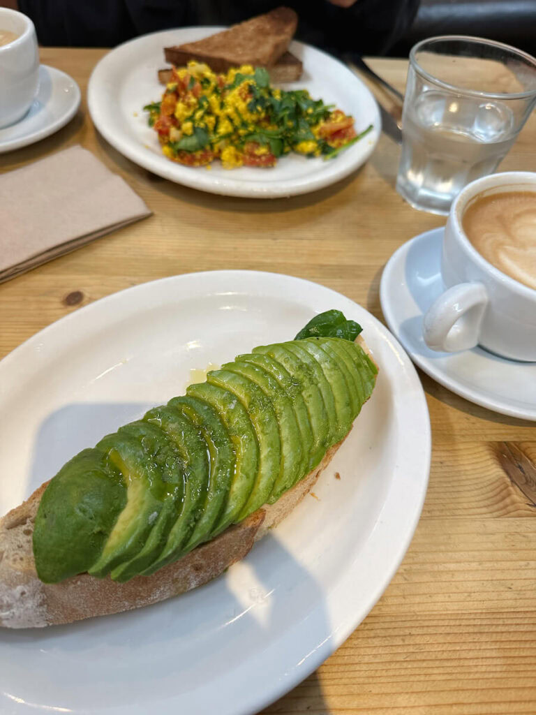 An avocado toast next to a latte in a white mug at Carmel Belle. There is a tofu scramble in the background. 