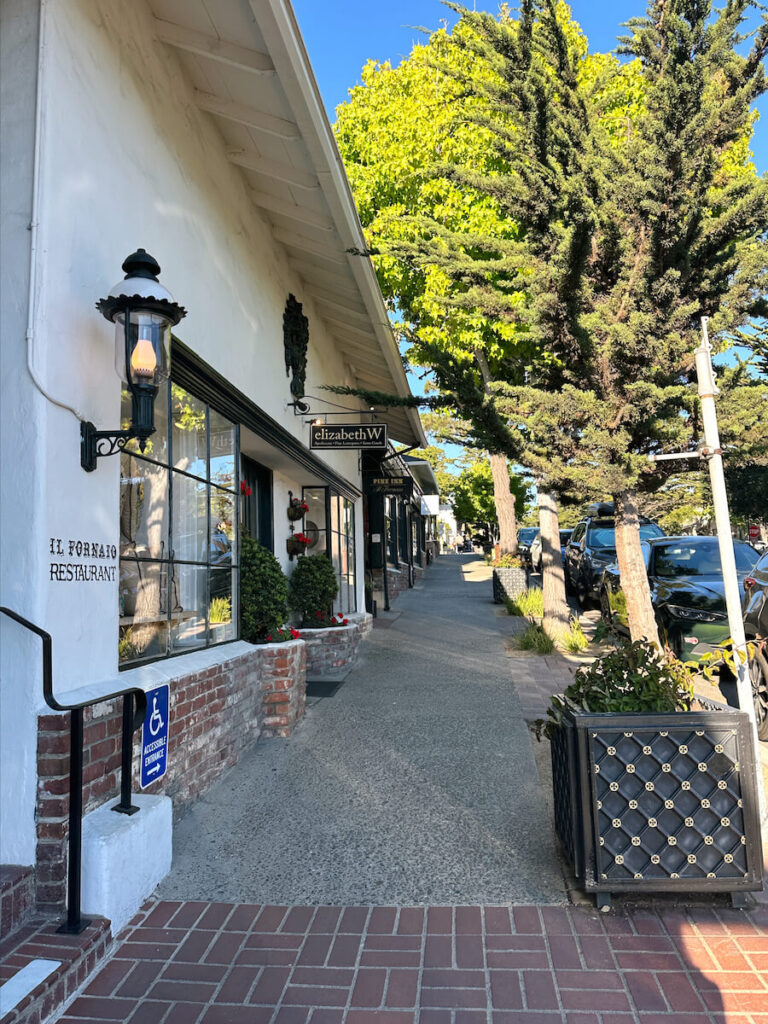 A tree lined sidewalk on Ocean Avenue. There are storefronts on the right, an an oil lamp on the building.