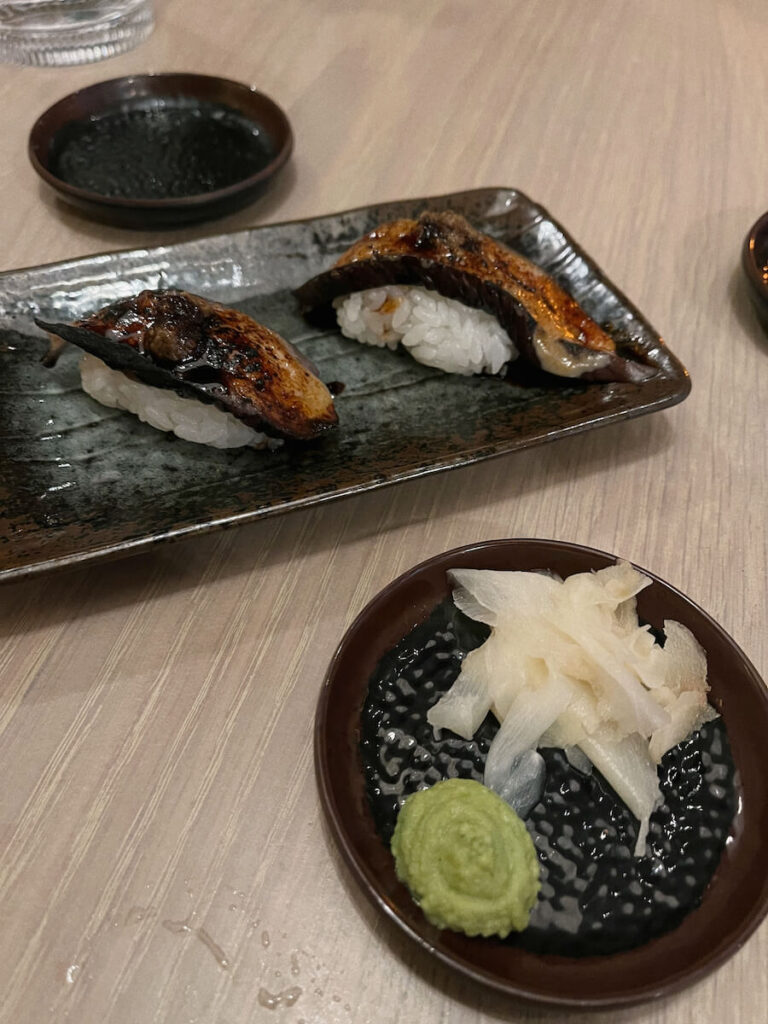 Eggplant nigiri on a dark plate, with a round small plate of ginger and wasabi in front