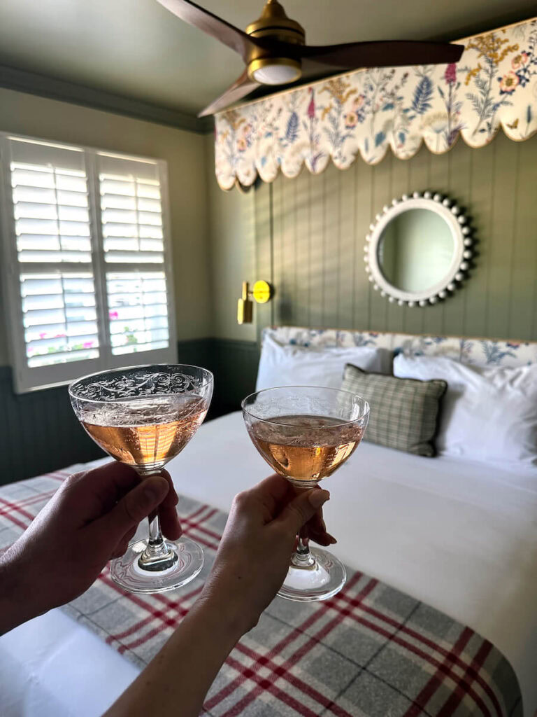 Two hands holding glasses of rose in front of a bed