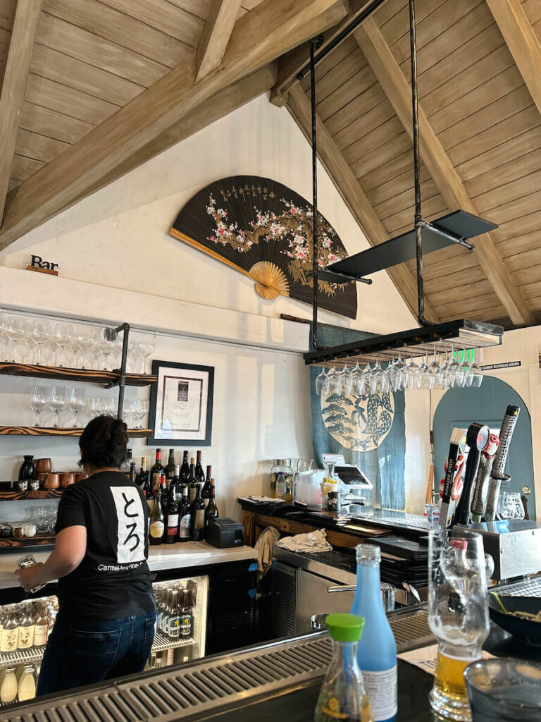 The interior of Toro Sushi. There is a large decorative fan on the wall with pink floral detailing, and the sushi bar is in the foreground.