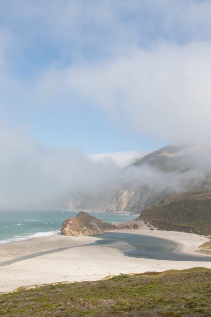 Fog rolling out the the sea above a large rock on a beach in Big Sur