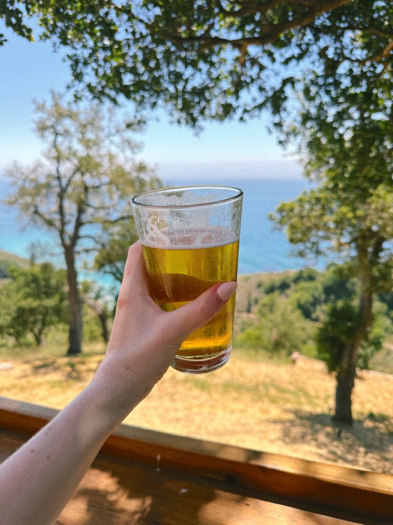 A hand holding a beer at Nepenthe. The view of trees in front of the ocean is in the background