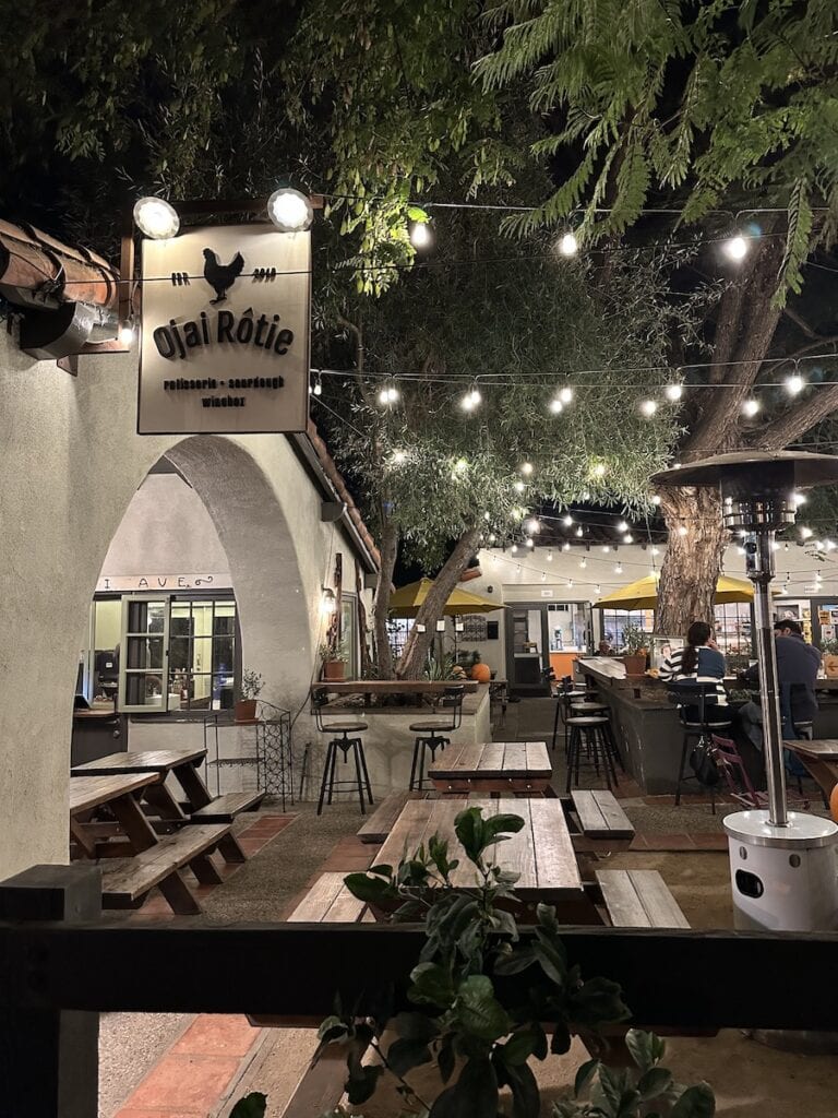 A sign for Ojai Rotie over multiple picnic tables. There are string lights above the tables. 