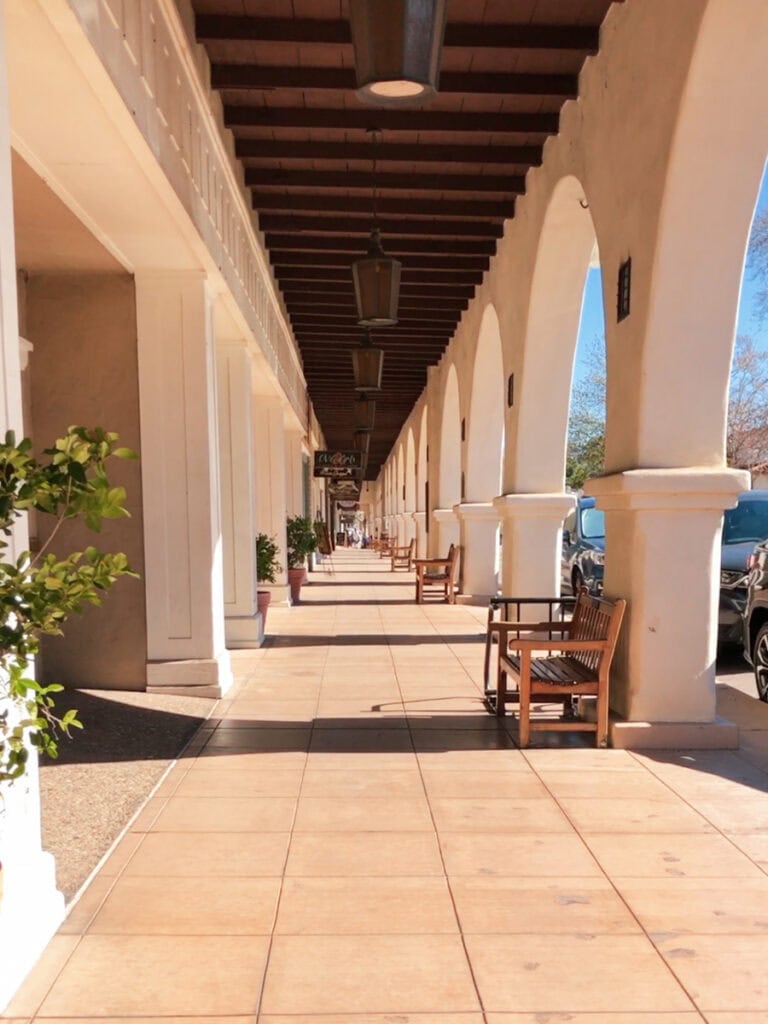 A covered pedestrian walkway in Downtown Ojai. There are multiple arches on the right side, and benches lining the walkway