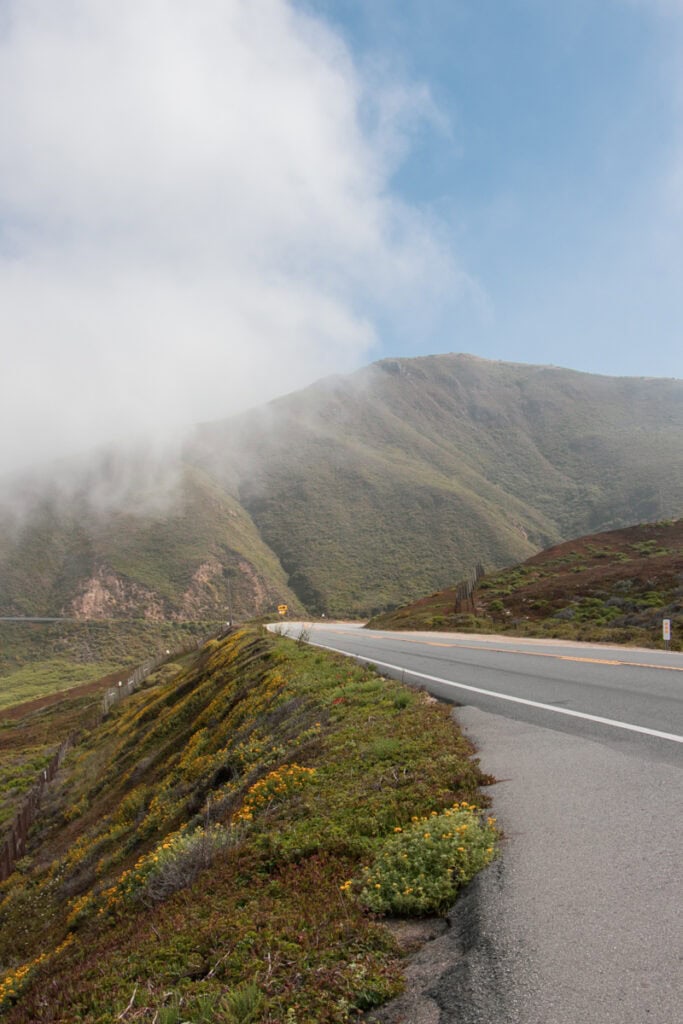 The Pacific Coast Highway in the foreground, with mountains in the background. There is some thin fog over the mountains, and you can see blue sky behind it. 