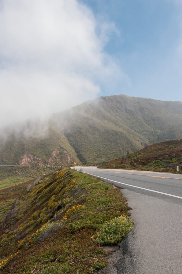 The Pacific Coast Highway in the foreground, with mountains in the background. There is some thin fog over the mountains, and you can see blue sky behind it.