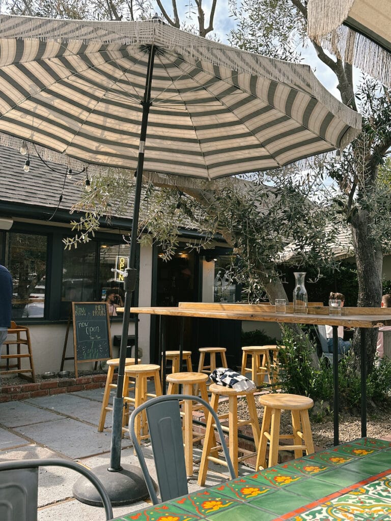 The patio outside of Three Birds. There are wooden stools around a bar next to an olive tree, and a striped umbrella. 