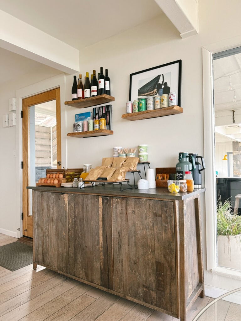 A counter with breakfast items (including fruit, breads, and orange juice) at Cambria Beach Lodge. There are shelves with alcoholic drinks above this counter. 