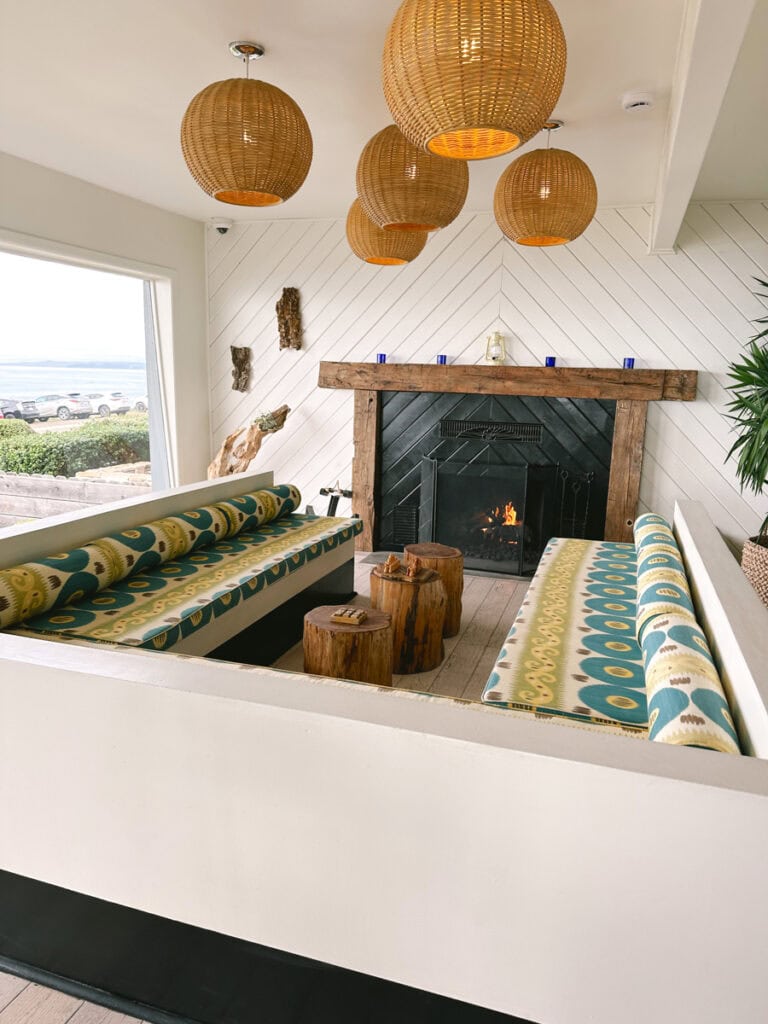 A wraparound couch with green and blue patterned cushions in front of a fireplace at Cambria Beach Lodge. There are circular rattan-style lamps hanging from the ceiling.