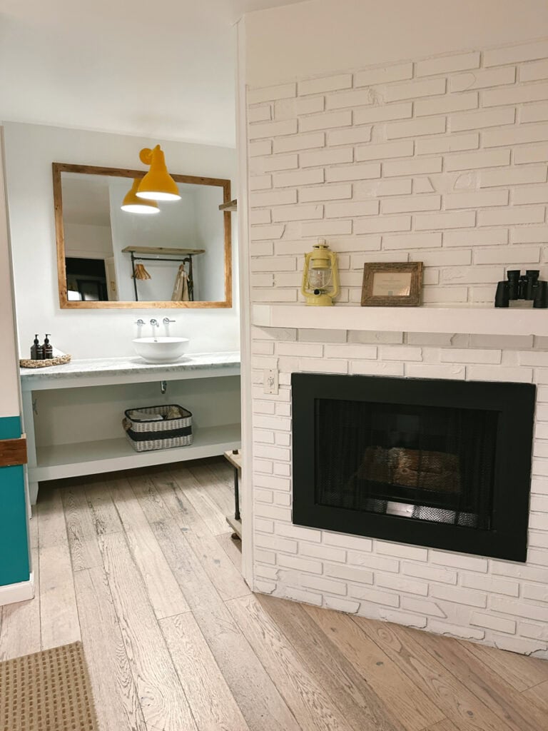 A small white brick fireplace and a hotel room sink and mirror behind it. 