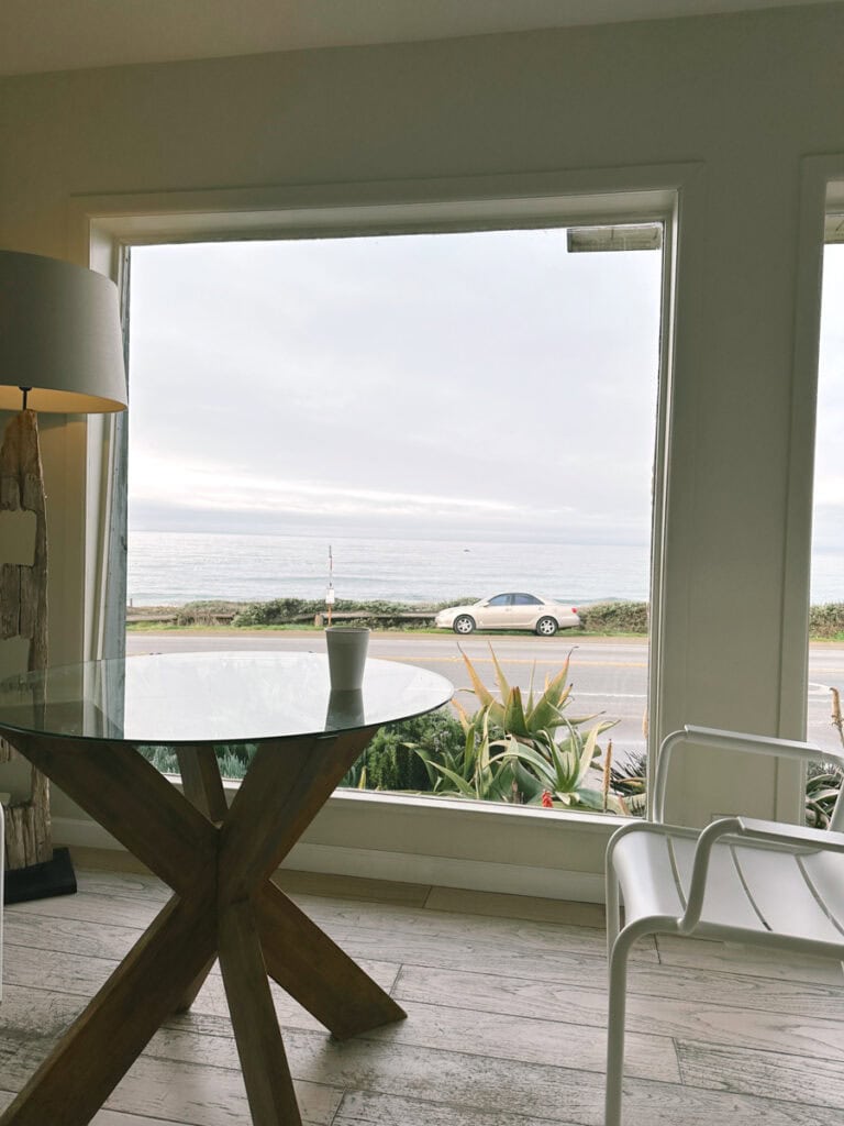 A glass table and white chair in front of a large window looking out to the ocean. 