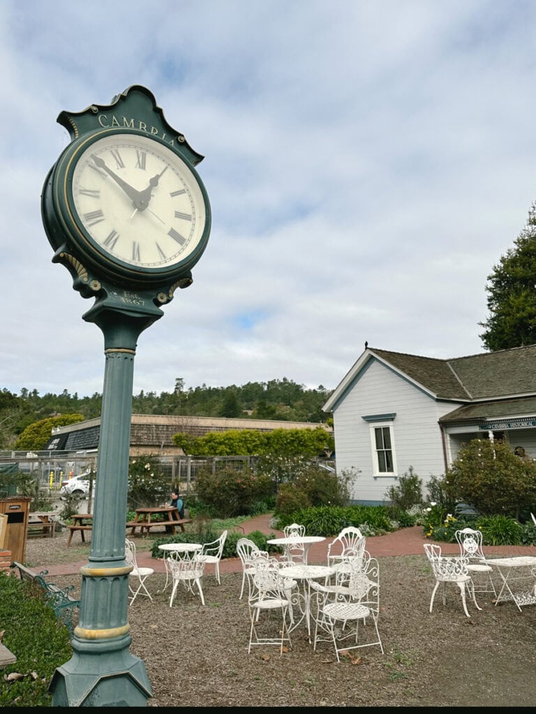 A green town clock in front of an outdoor space with white tables and chairs