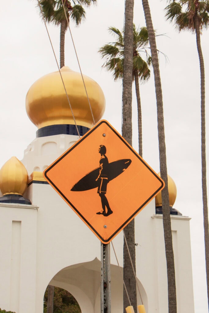 A yellow street sign with a man holding a surfboard. There is a temple with gold domes in the background, as well as palm trees