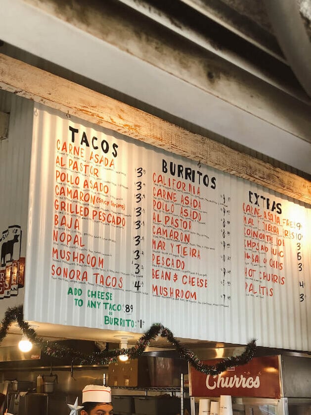 A woman ordering at the food counter at The Taco Stand. A large menu painted on corrugated metal is above the register.