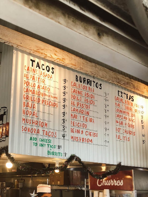 A woman ordering at the food counter at The Taco Stand. A large menu painted on corrugated metal is above the register.