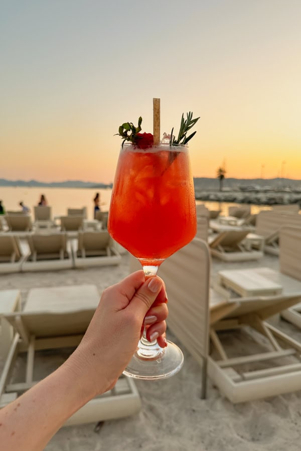 A handing holding a bright orange cocktail in front of the view of the sunset on the beach