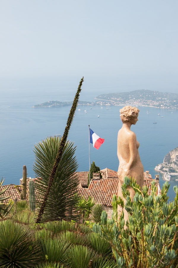 A statue of a woman surrounded by plants, and a French flag on the roof of a building in the background