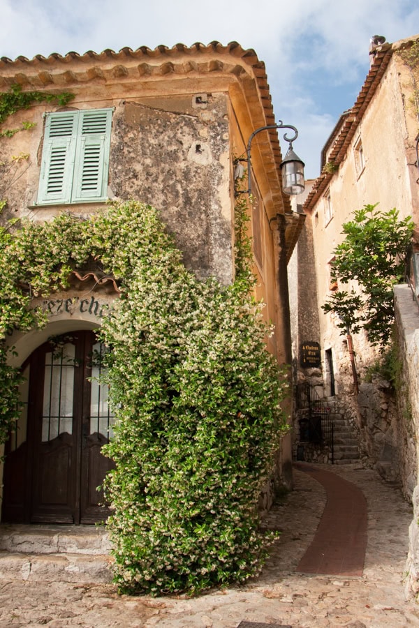 A stone building covered in greenery next to a curved path. There is a rounded wooden door at the entrance to the building.