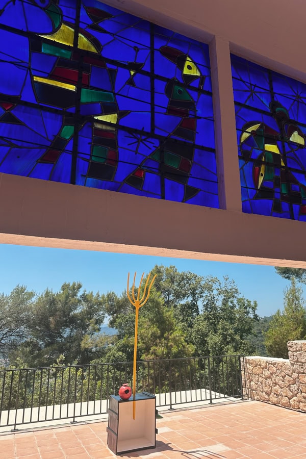 An overhang featuring blue stained glass in front of a yellow pitchfork-shaped sculpture in the outdoor gardens and Fondation Maeght