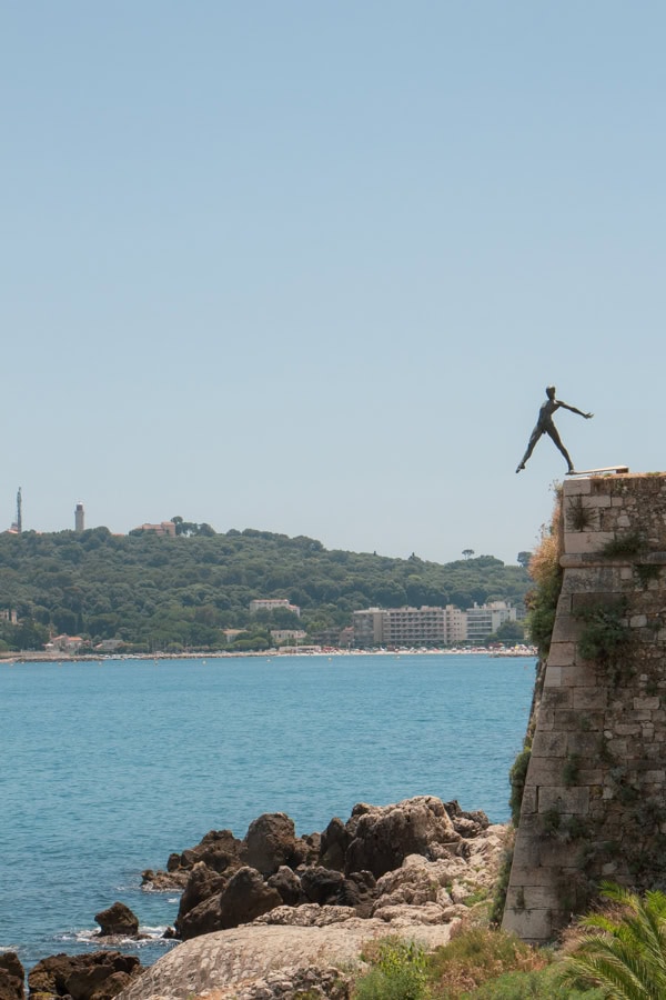 A metal statue of a person stepping off the ledge of a stone wall that overlooks the sea