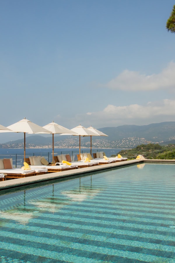 Sun loungers with white umbrellas over them in front of the pool. You can see the ocean in the background.