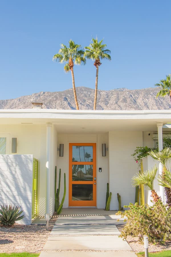 A white mid-century modern house with an orange door in Indian Canyons. There are desert plants in the front yard, and palm trees behind the house.
