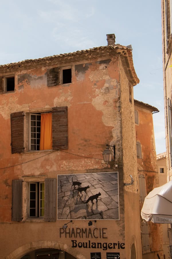 An old light orange colored building in Gordes with a mural of three cats on it.
