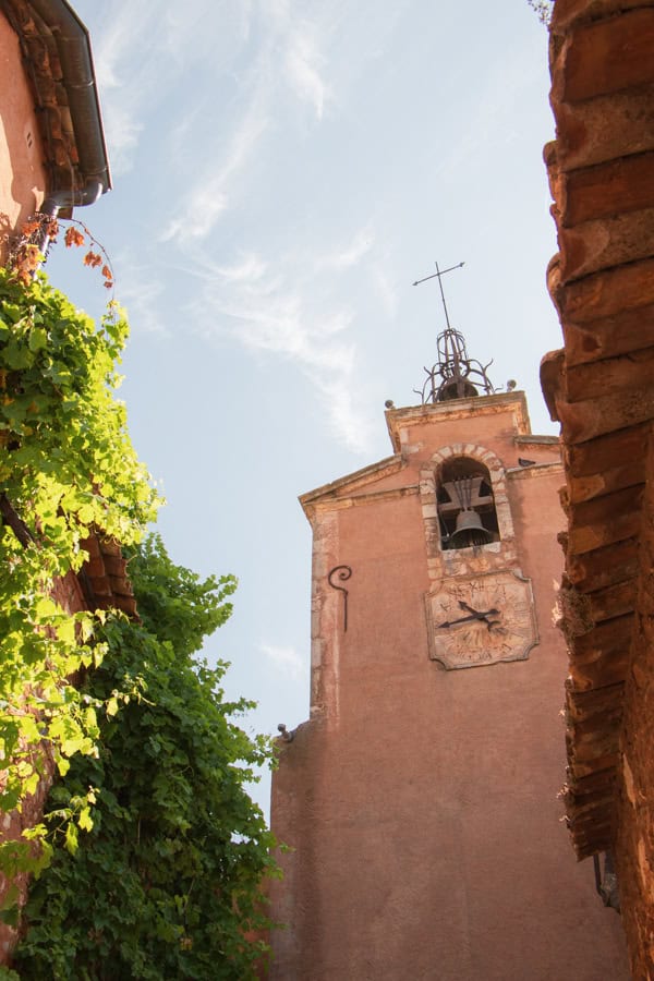 A pink-colored bell tower in Roussillon. There is greenery growing from the building nex to the bell tower