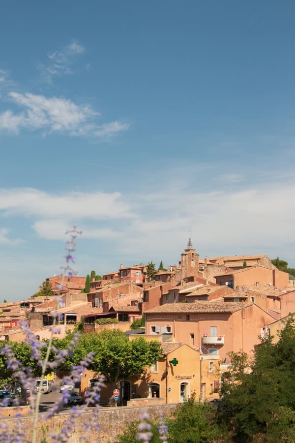A view of the red and orange-hued town of Roussillon from the distance. There are purple flowers in the foreground.