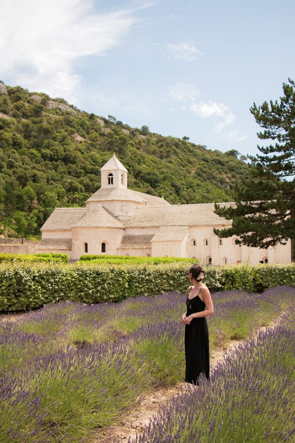 A woman in a black dress looking towards the Senanque Abbey. She is standing between rows of lavender in bloom.