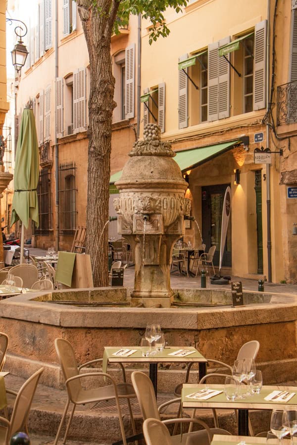 A fountain in the middle of a square in Aix. There are dining tables surrounding the fountain.