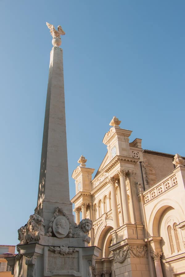 An ornate white statue in front of a larger ornate white building in Aix