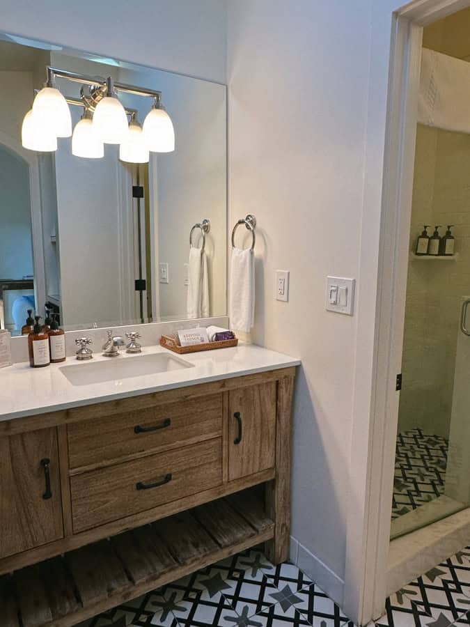 A hotel room bathroom at Hotel Ynez. There are three light pendants over the white vanity with wooden base, and there is a shower in the room next to the vanity.