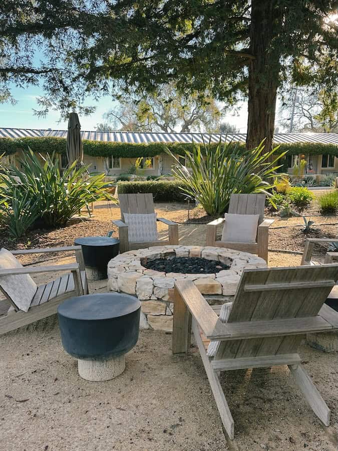 Five wooden lounge chairs around a round stone fire pit at Hotel Ynez. There is a large tree behind the chairs, as well as landscaping surrounding the seating area.