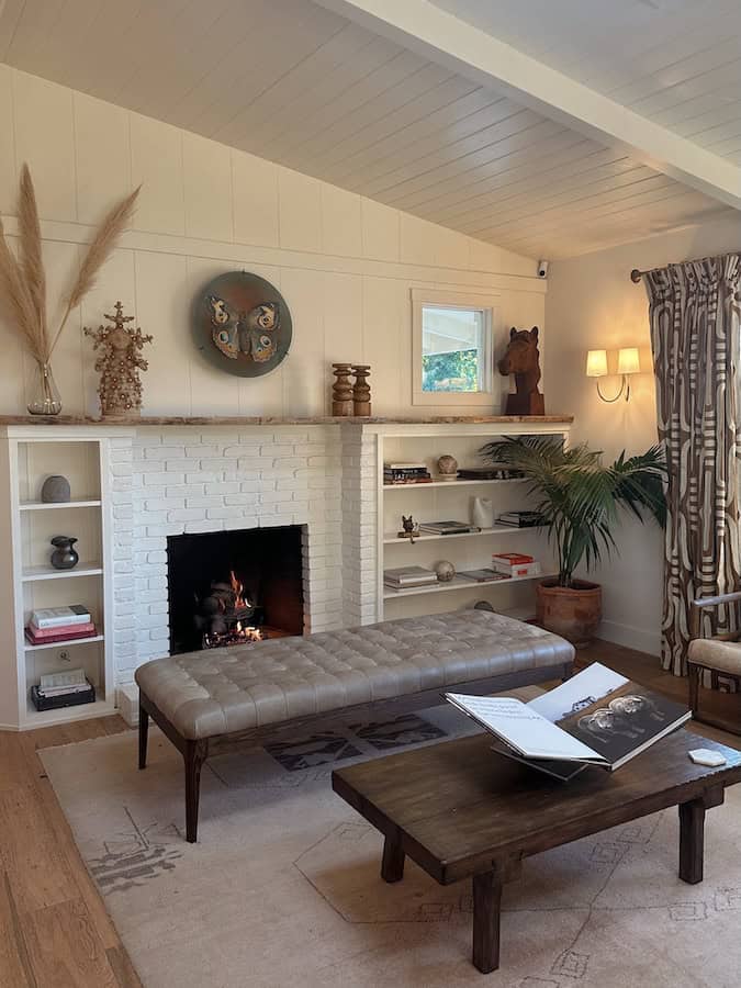 The indoor lounge at Hotel Ynez. There is a white stone fireplace with shelving on either side holding books and art pieces. There is a bench in front of the fireplace.