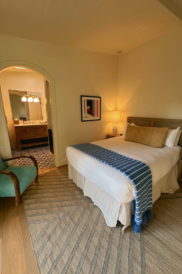 A hotel room at Hotel Ynez. There is a bed with a blue blanket and long yellow pillow, and a teal chair in the corner